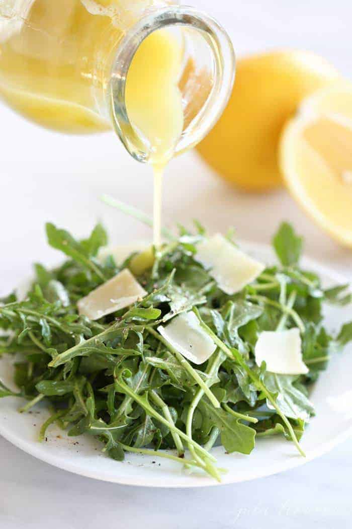 An arugula salad on a white plate, homemade vinaigrette pouring over the top.