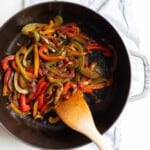 A cast iron pan filled with sauteed peppers, wooden spoon sticking out one side.