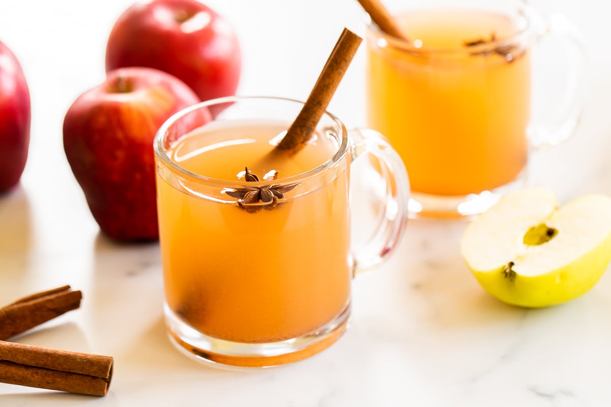 A clear glass mug full of homemade apple cider, with apples in the background.