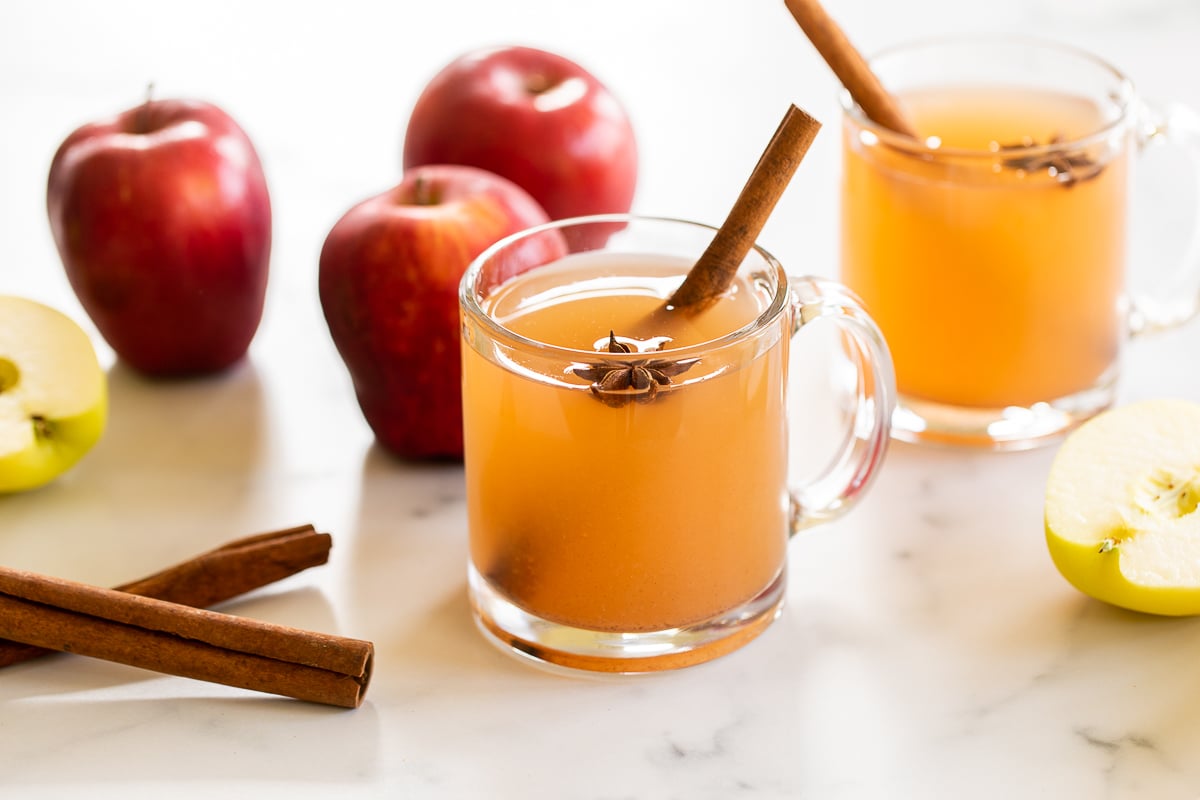 A clear glass mug full of homemade apple cider, with apples in the background.