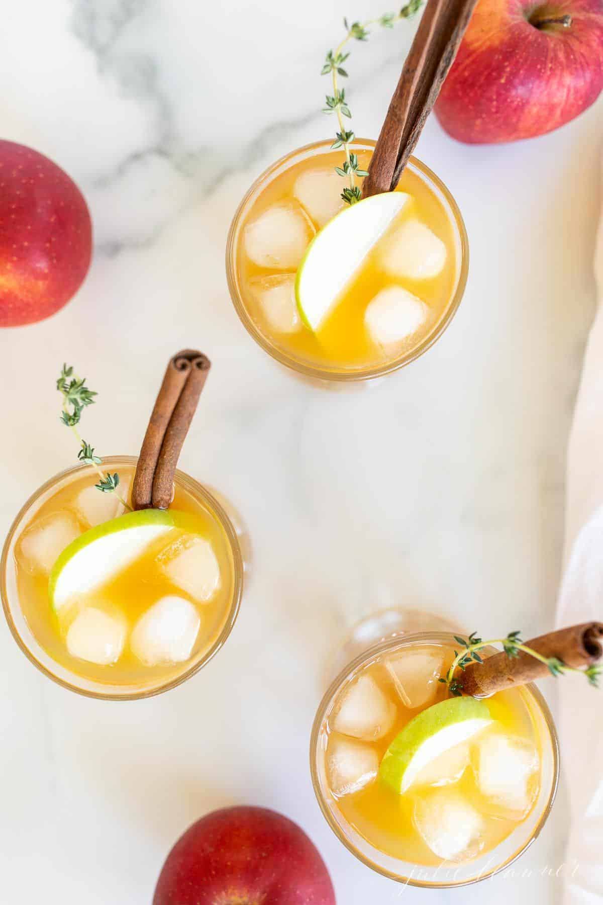Cocktail glasses filled with an apple cider cocktail on ice, garnished with thyme, cinnamon stick and apple slice, apples to the side.