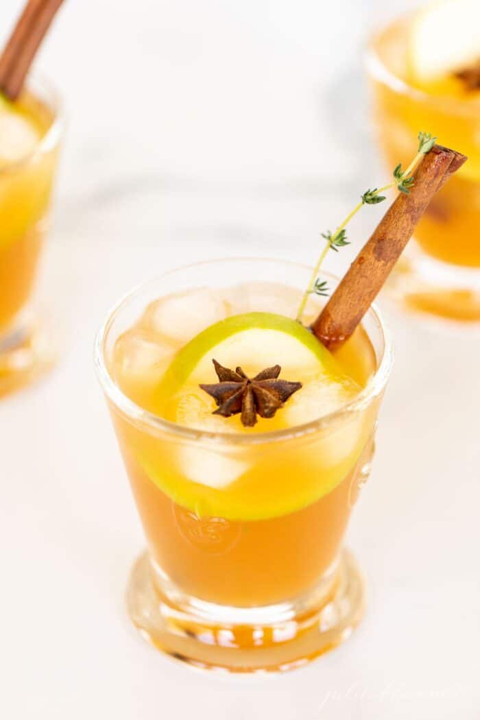 A cocktail glass filled with an apple cider cocktail on ice, garnished with thyme, cinnamon stick and apple slice.