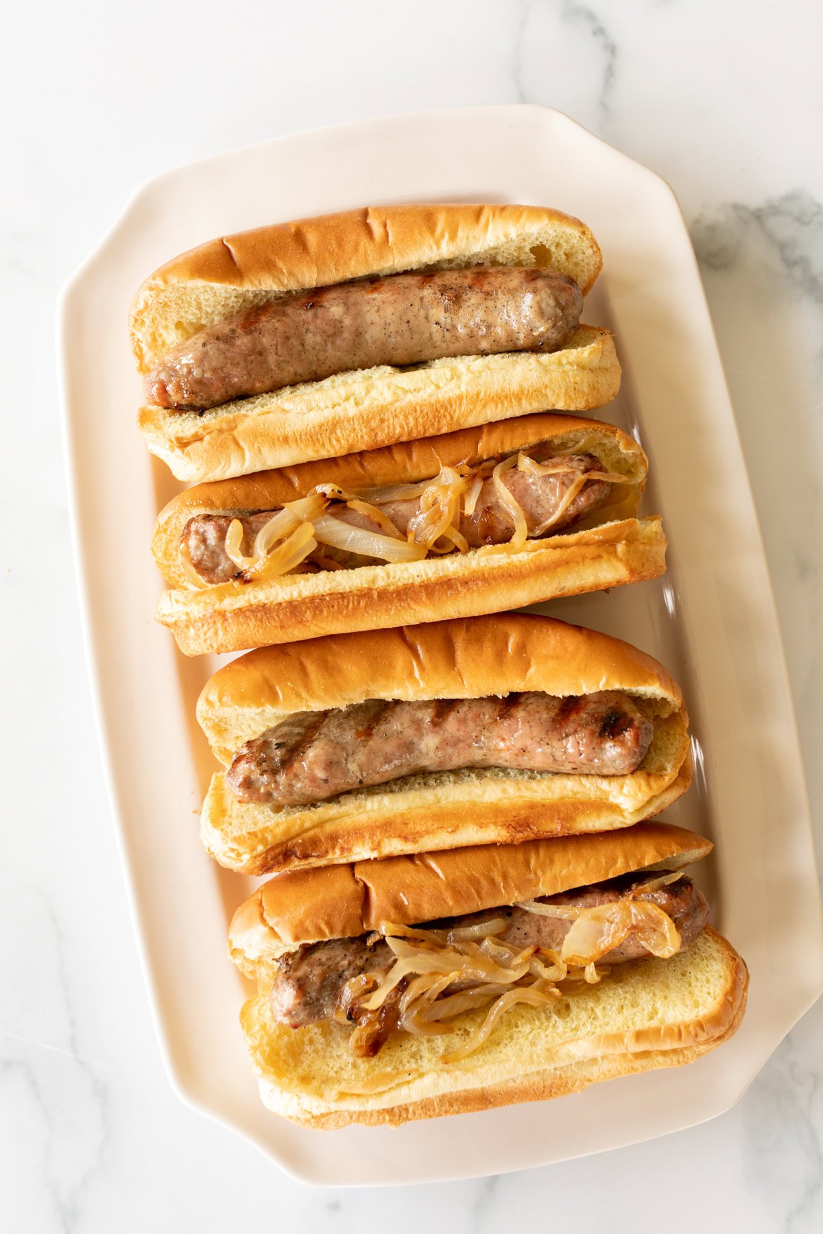Four bratwursts in buns topped with cooked onions are arranged on a rectangular white plate on a marble surface.