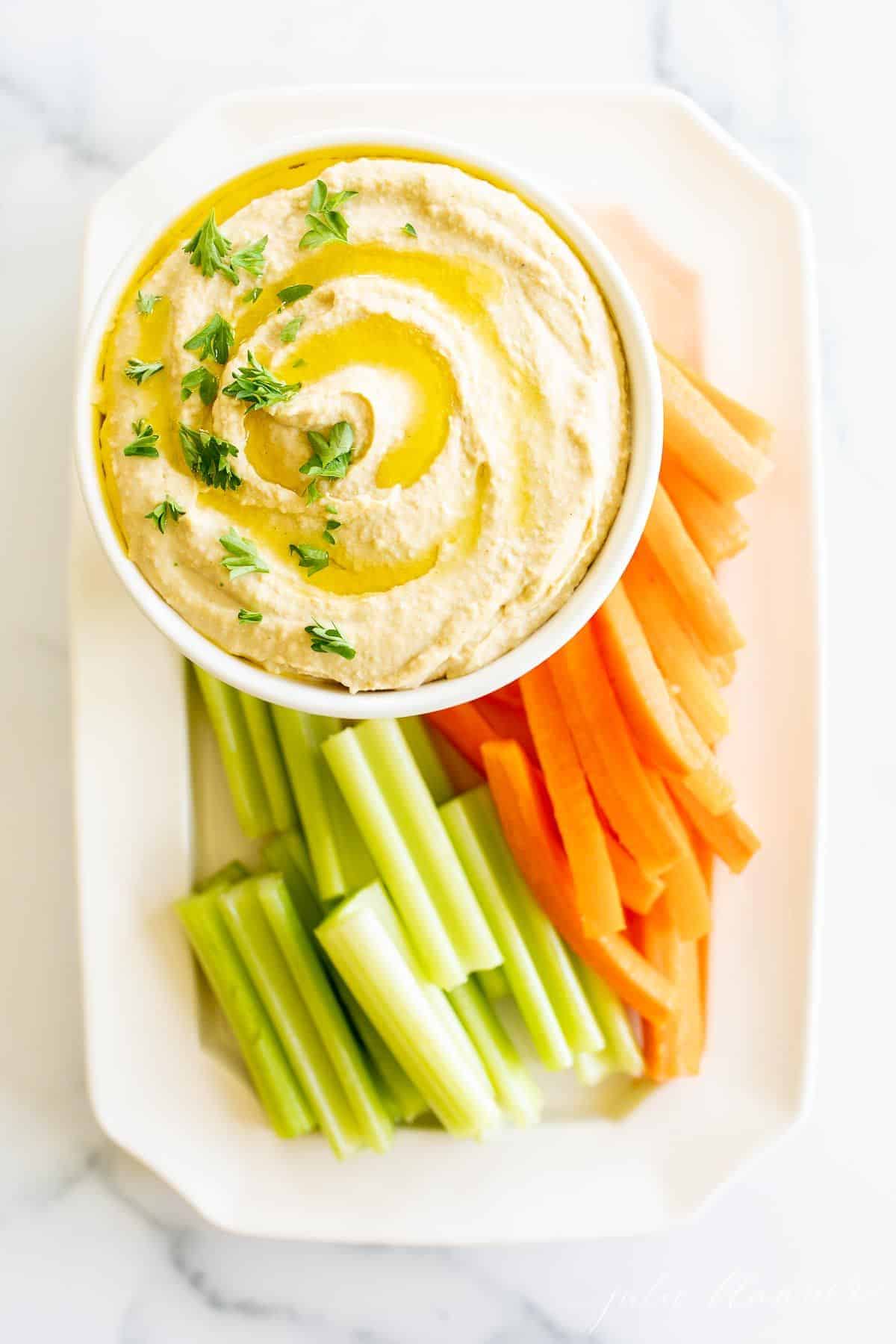 A bowl full of homemade hummus sitting on a platter of vegetables for dipping.