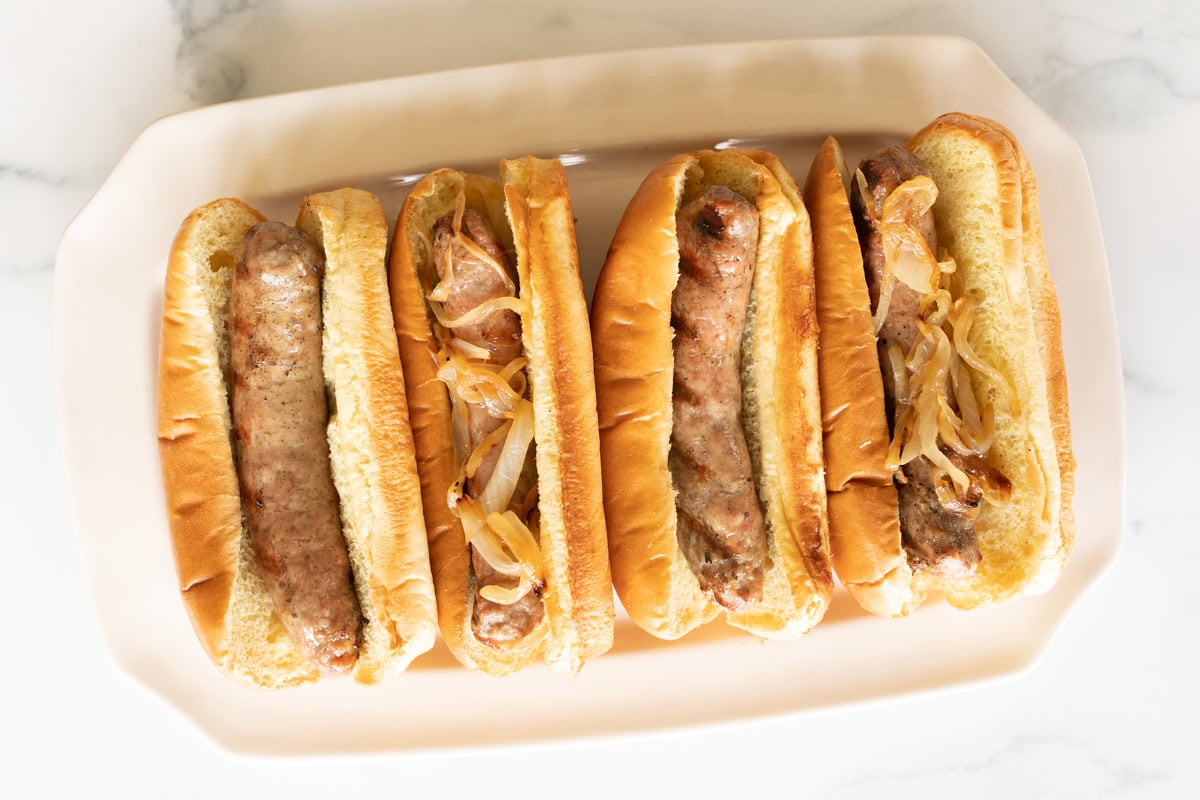 A platter holding four bratwurst sausages in buns topped with cooked onions, arranged in a row on a white surface.