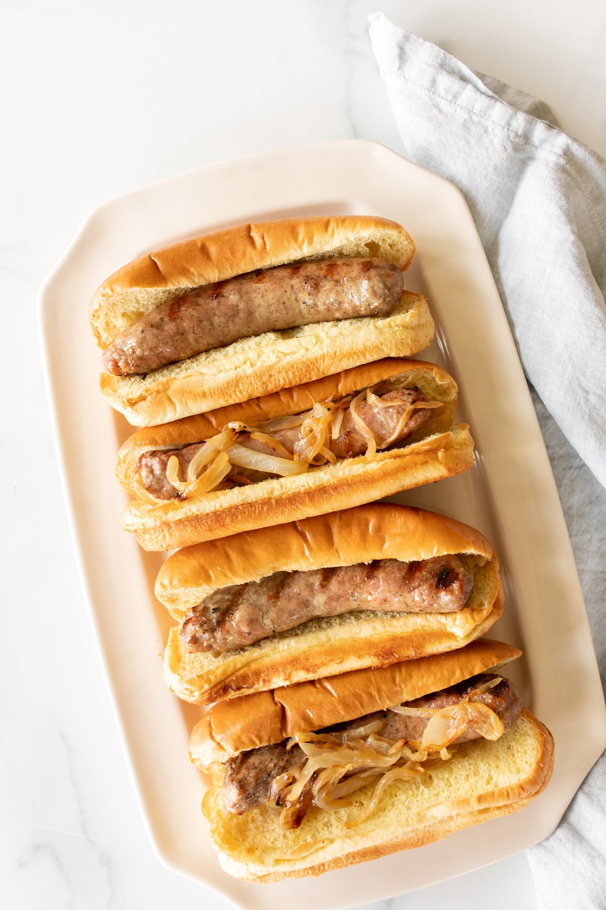 A white plate holding four cooking bratwursts in buns with caramelized onions, placed next to a light gray cloth napkin on a marble surface.