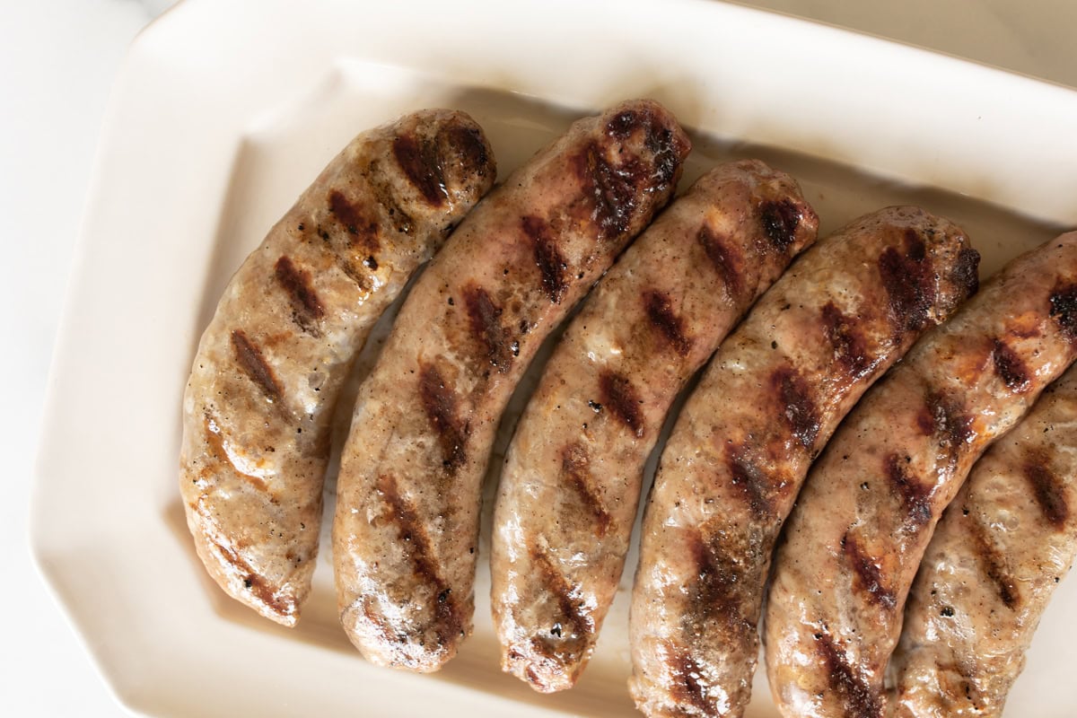 Grilled bratwurst sausages with perfect grill marks, arranged in a row on a rectangular white plate.