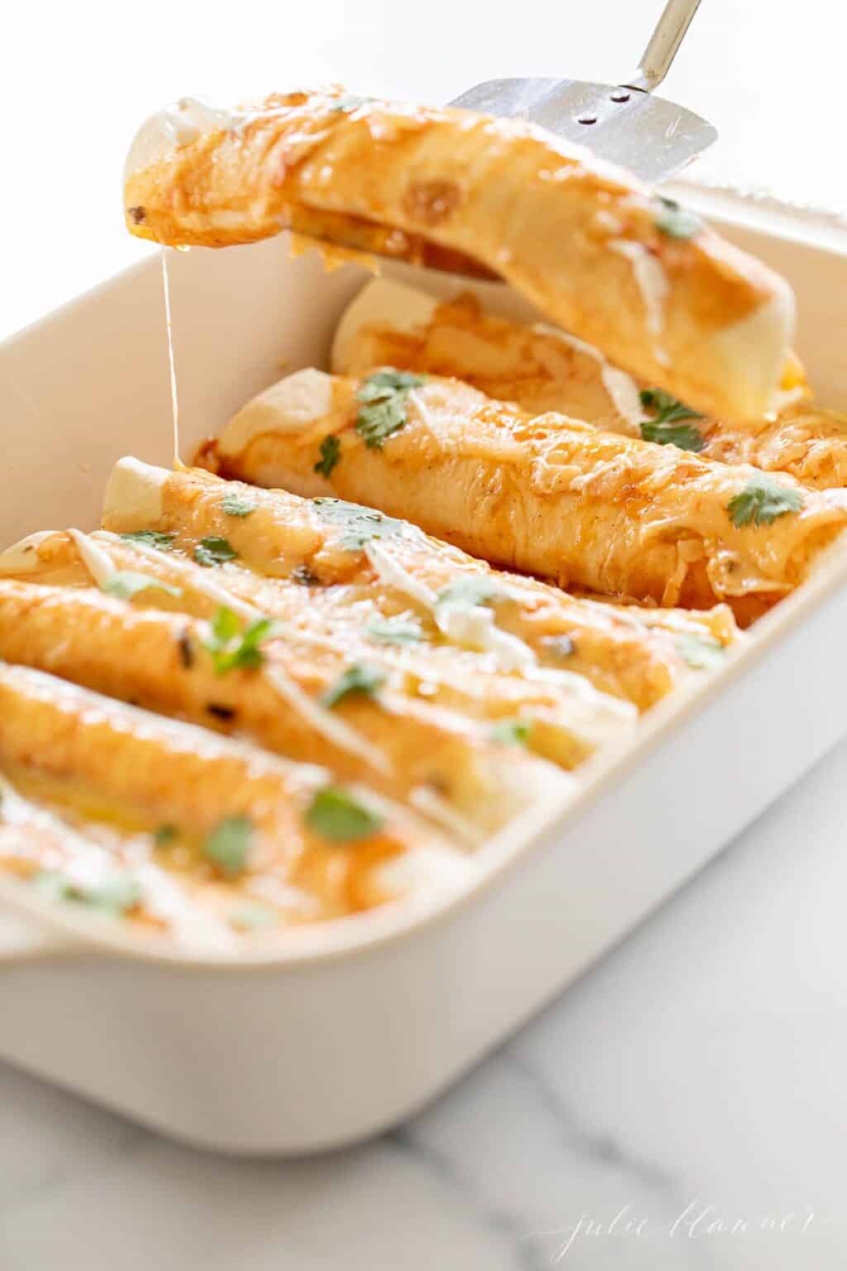 Cheese enchiladas in a white baking dish, a silver spatula lifting a serving out. 