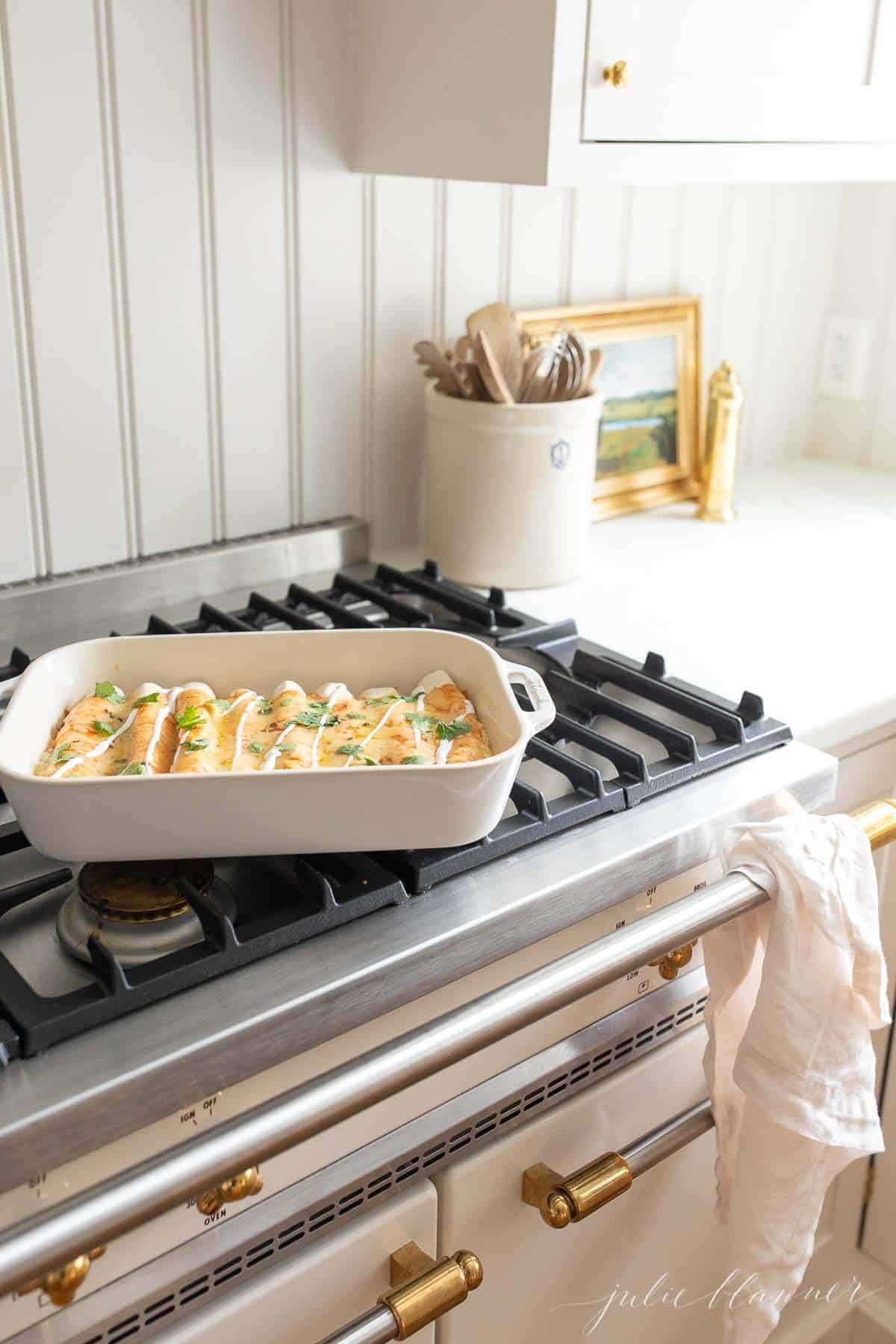 A white casserole dish full of cheese enchiladas sitting on top of the stove in a white kitchen.