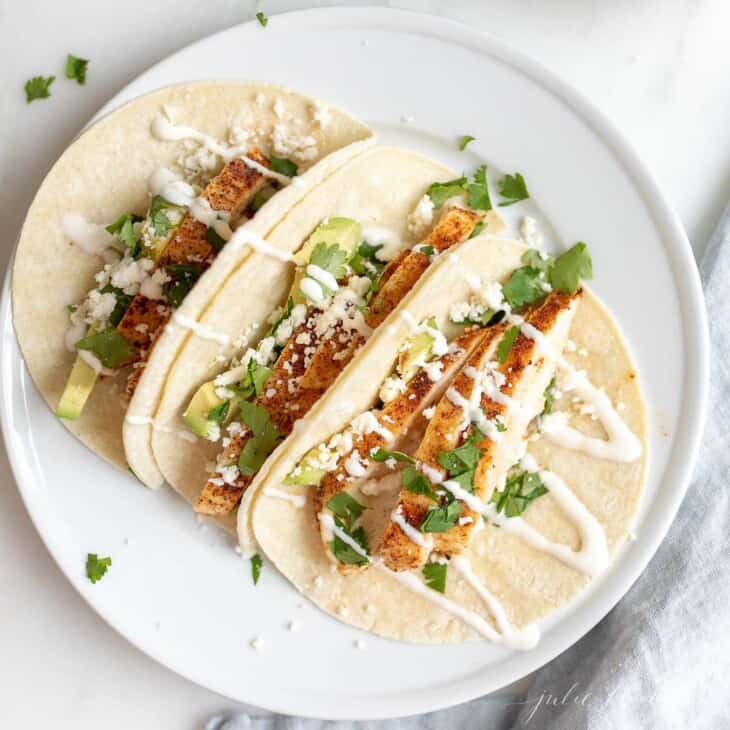 A white plate full of soft chicken tacos drizzled with crema.