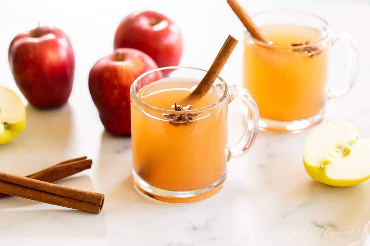 A marble surface with clear mugs full of homemade apple cider, whole apples and cinnamon sticks to the side.
