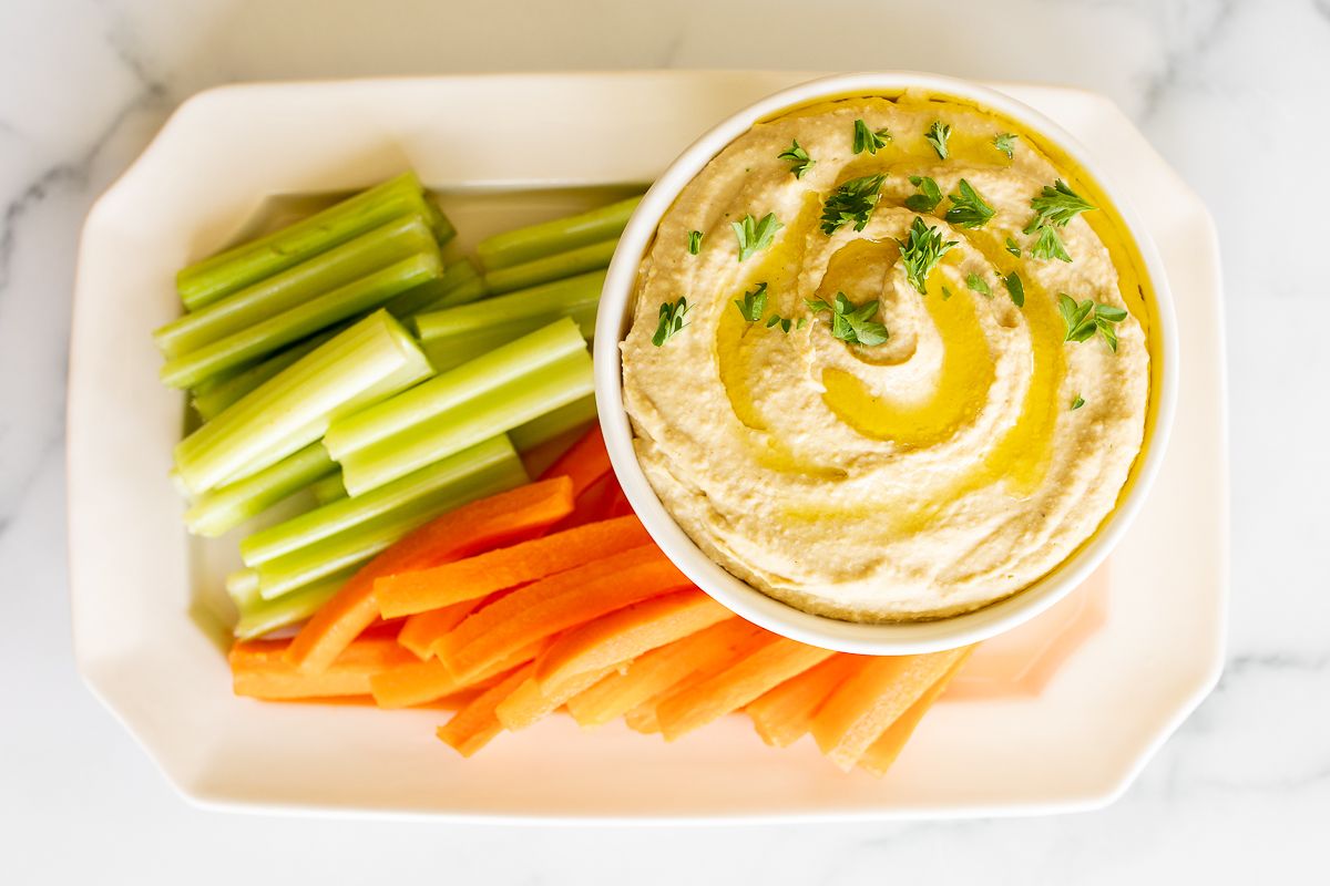 A white bowl of hummus surrounded by a platter of vegetables for dipping.