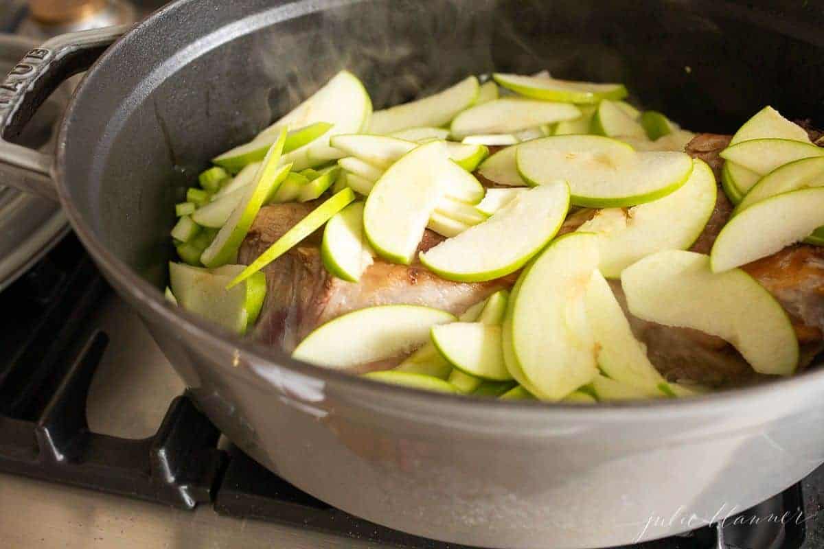 A cast iron pot on a stovetop with pork covered in sliced apples for a pork ragu recipe.