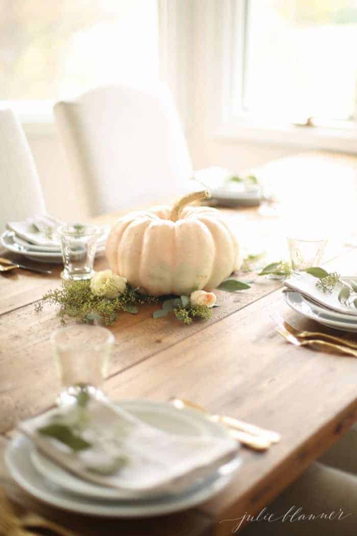 A wooden farm table set for a fall party with a white pumpkin and fresh flowers as a fall centerpiece.
