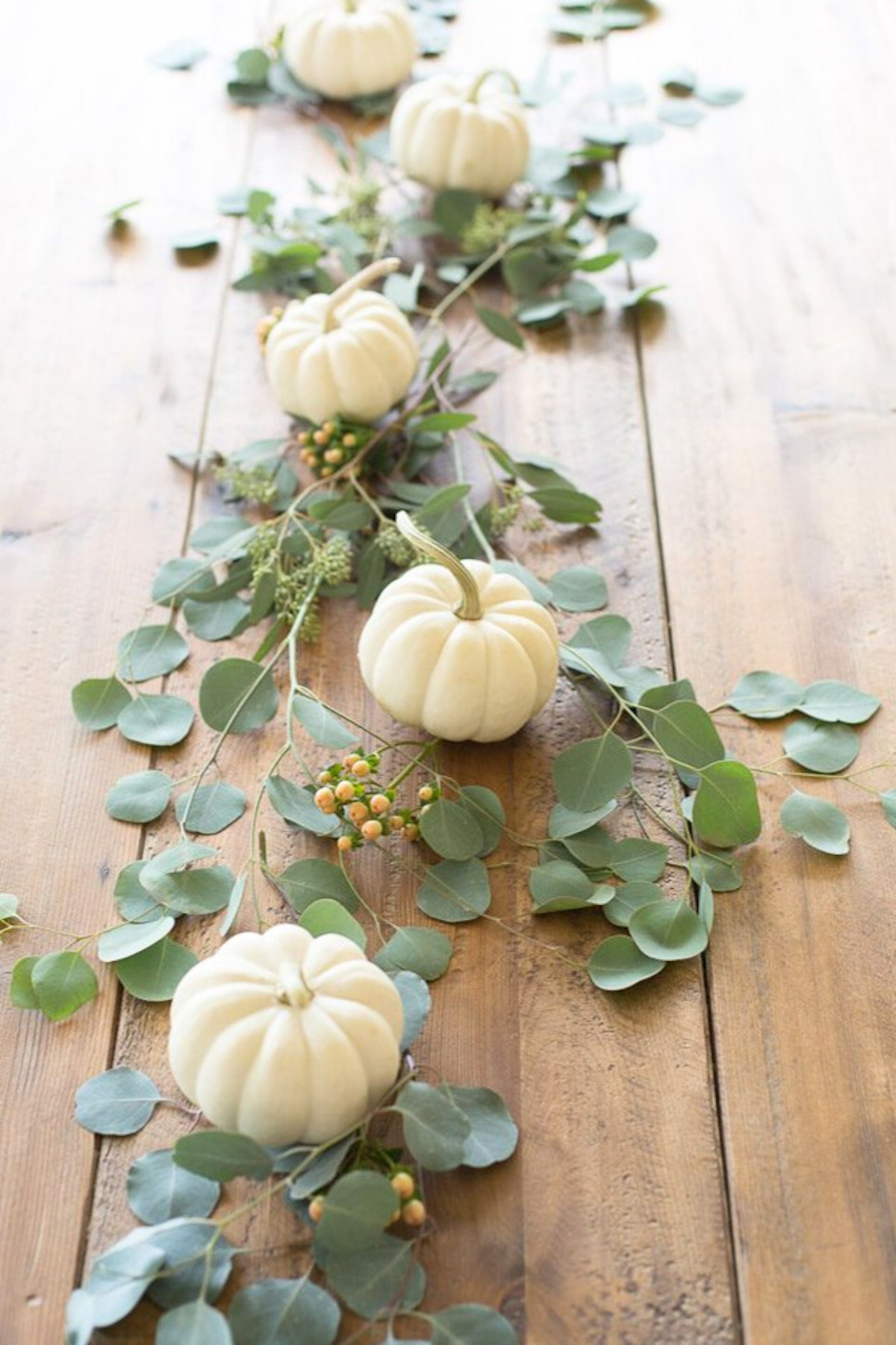A fall centerpiece of mini white pumpkins and eucalyptus down the center of a wood table.