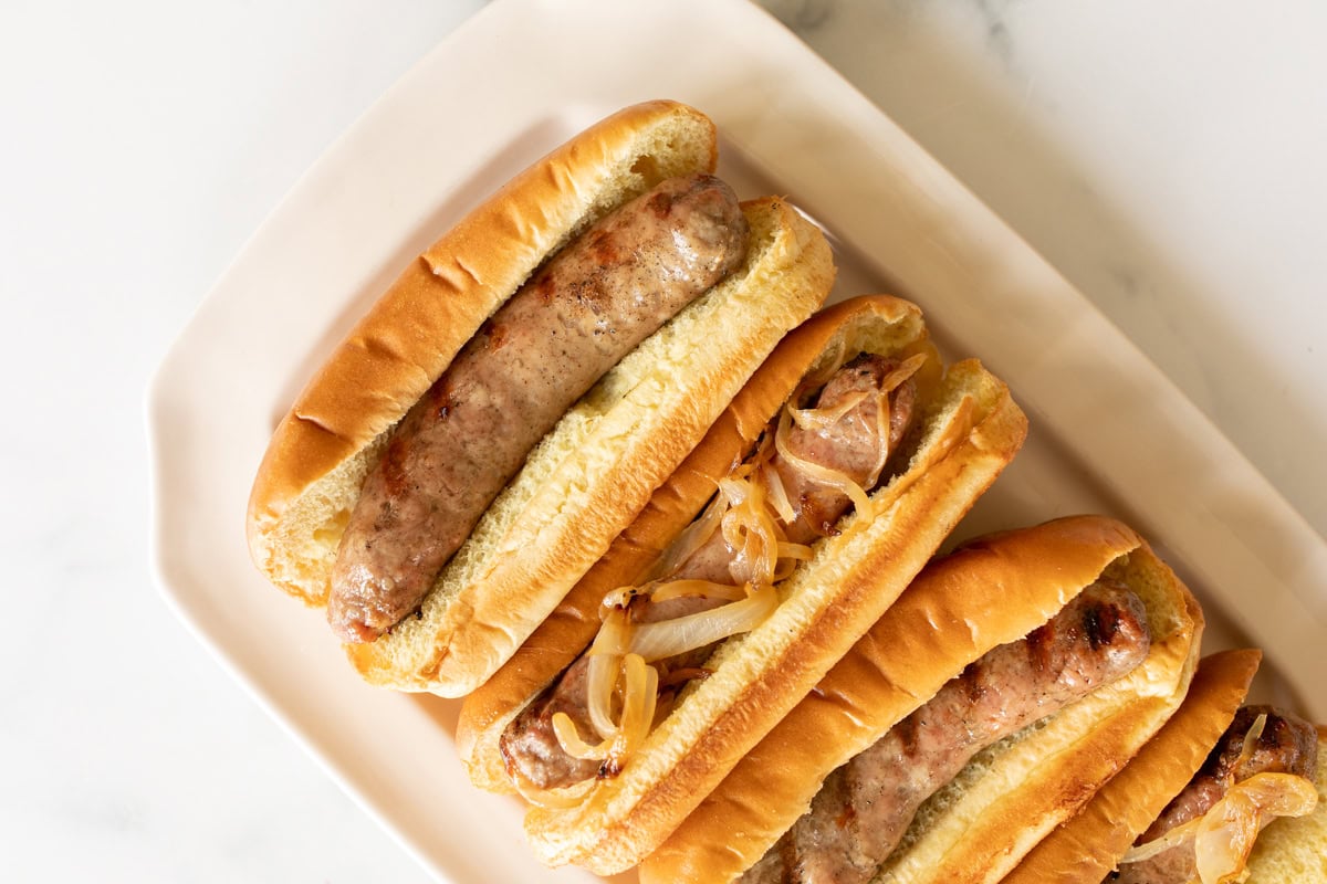 Three expertly grilled bratwursts topped with sautéed onions in hot dog buns, all thoughtfully arranged on a pristine white plate.