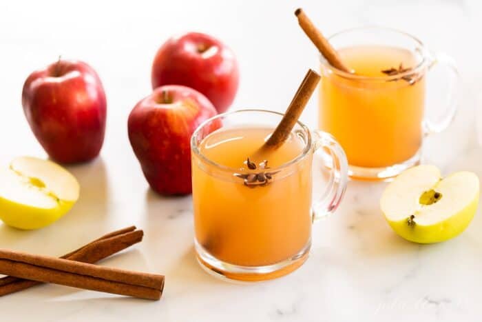 A marble surface with clear mugs full of homemade apple cider, whole apples and cinnamon sticks to the side.