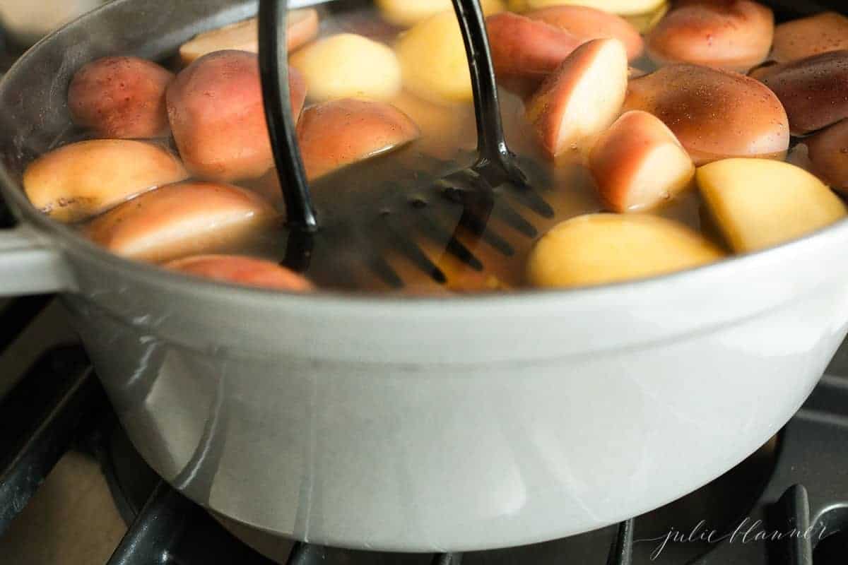 Close up shot of sliced apples in a pot of water, boiling into a homemade apple cider recipe, a potato masher in the pot.