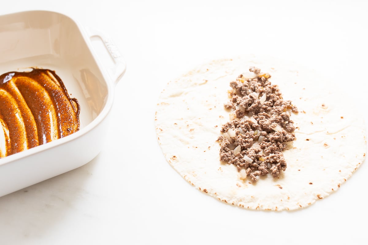 A tortilla topped with beef, next to a pan full of enchilada sauce