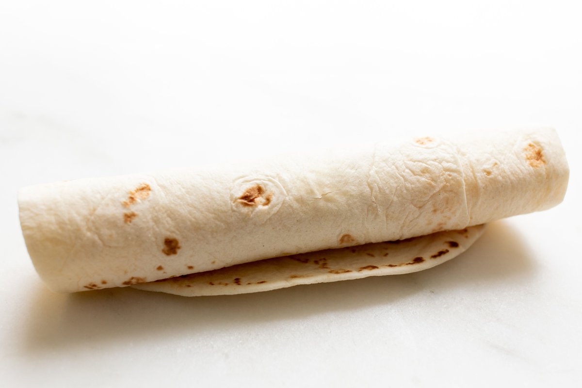 A flour tortilla rolled into an enchilada shape, on a marble countertop.