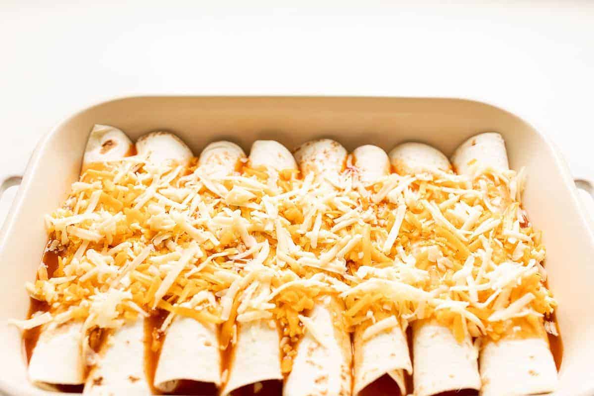 A white casserole dish filled with rolled tortillas covered in enchilada sauce and cheese to make cheese enchiladas.