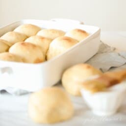 A white baking pan full of a homemade dinner roll recipe, two rolls in foreground with a bowl of melted butter for brushing across the top.