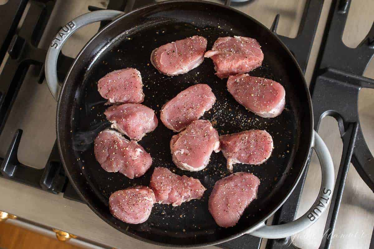 A cast iron pan on a stove top full of sliced pork medallions seasoned with a little salt and pepper.