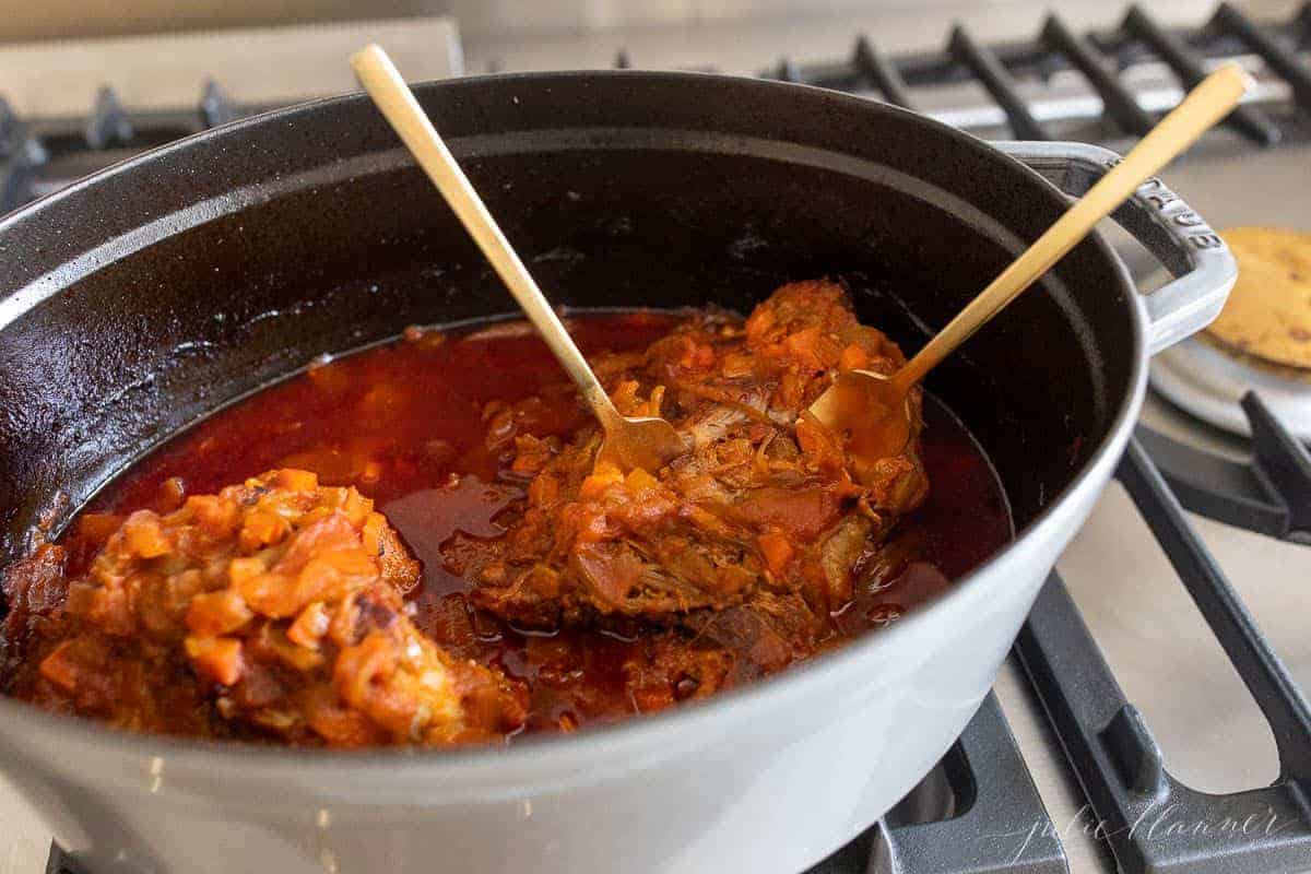 A pork shoulder surrounded by cooked apples and veggies in a cast iron pot, two forks inside to pull the pork.