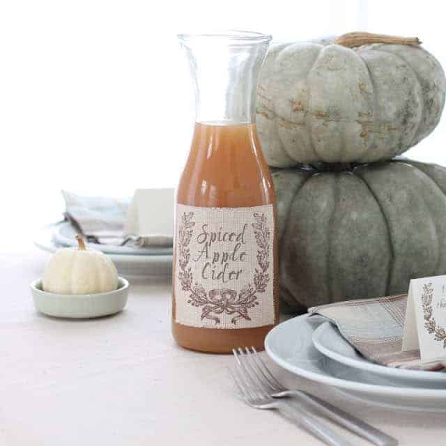 jar filled with apple cider with printed label on table setting, pumpkins stacked in the background.