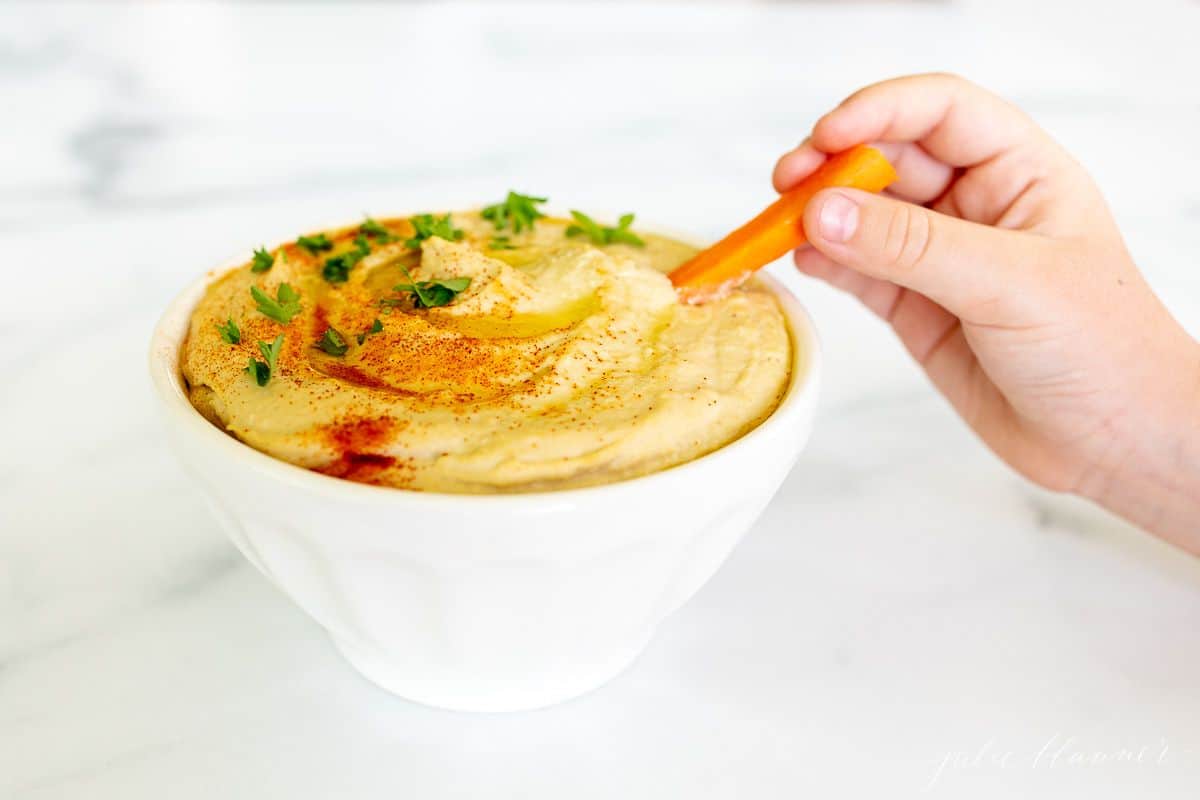 A hand reaching into a white bowl full of homemade hummus with a carrot stick.
