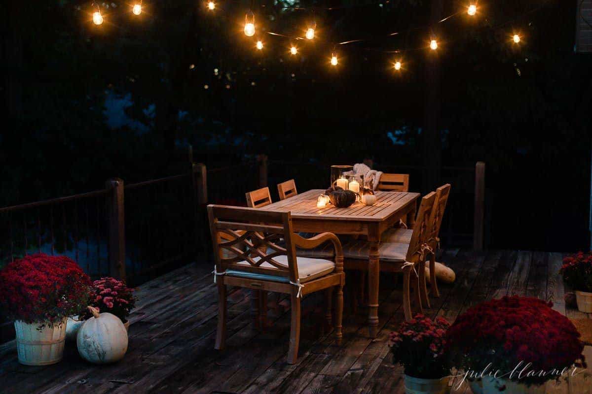 A deck dining area decorated for fall with string lights, pumpkins and mums
