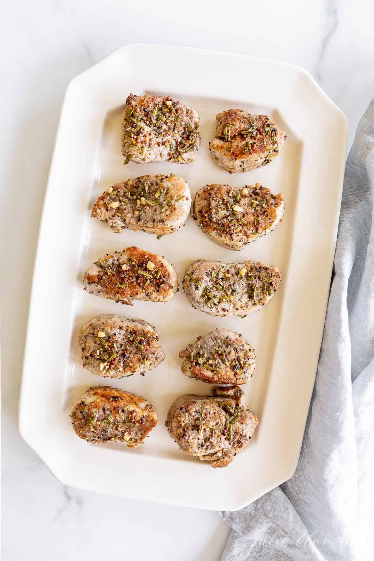 A white tray full of pork medallions topped with seasoning.
