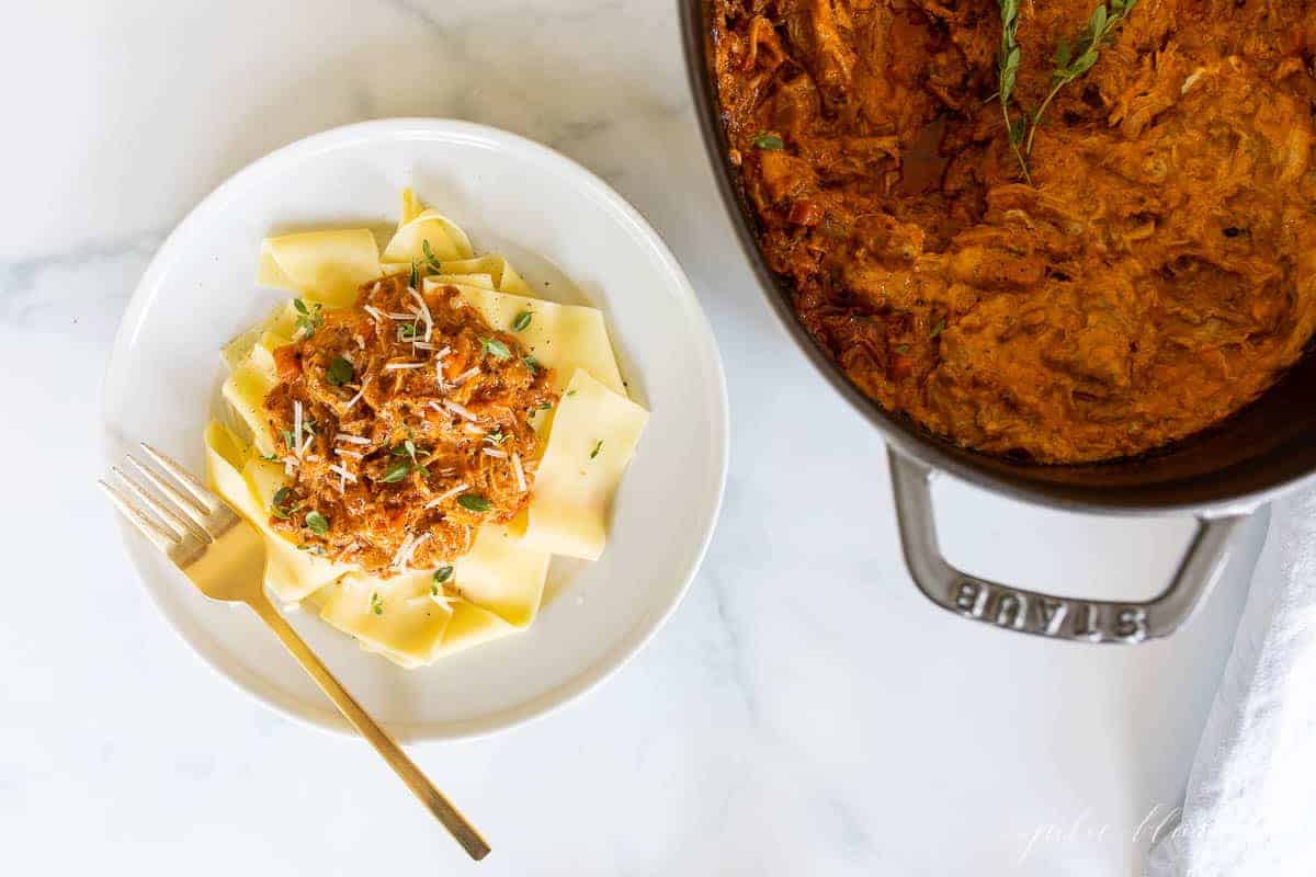 pork ragu on bed of pappardelle on white plate with gold fork next to cocotte