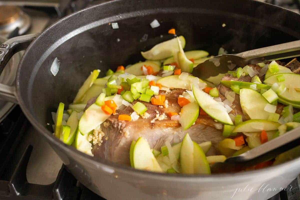 A cast iron pot on a stovetop with pork covered in sliced apples for a pork ragu recipe.