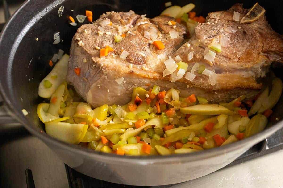 A pork shoulder surrounded by cooked apples and veggies in a cast iron pot.