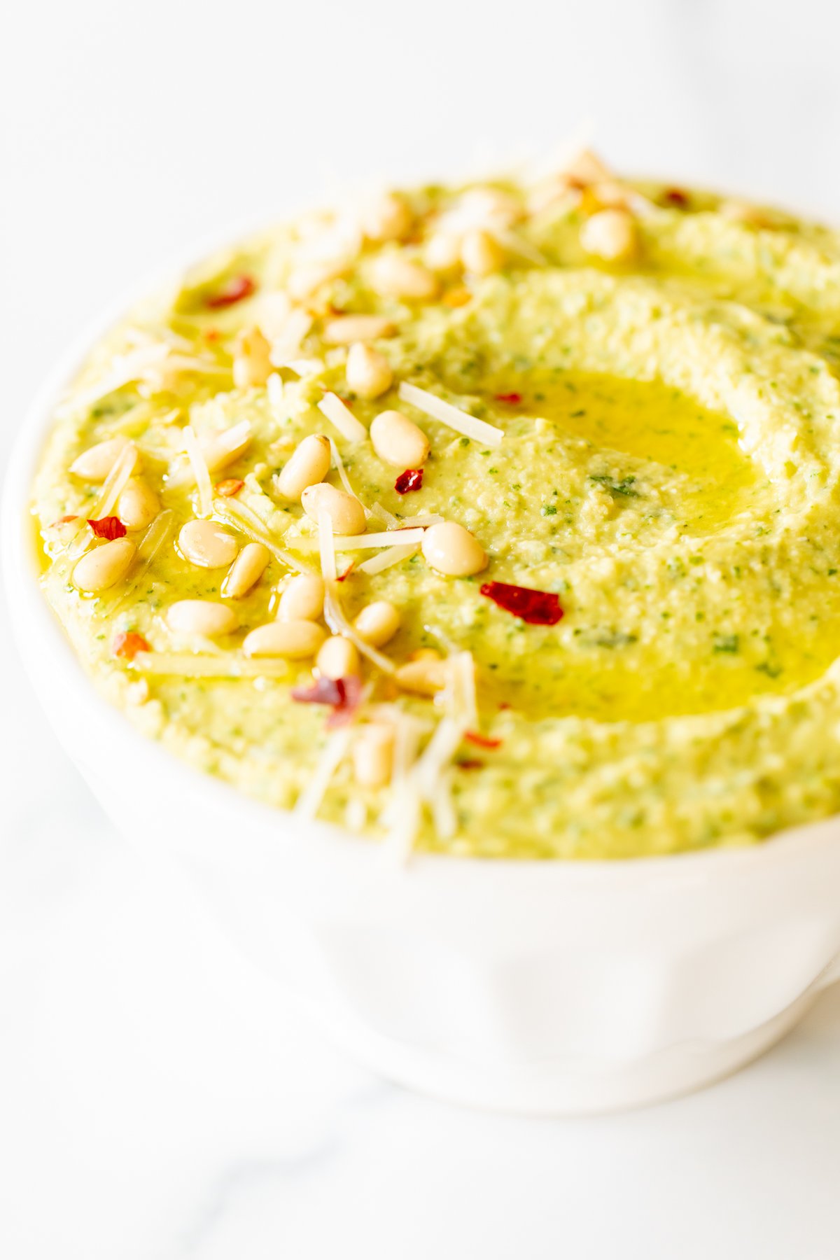 A white bowl full of basil pesto hummus, topped with pine nuts and parmesan.