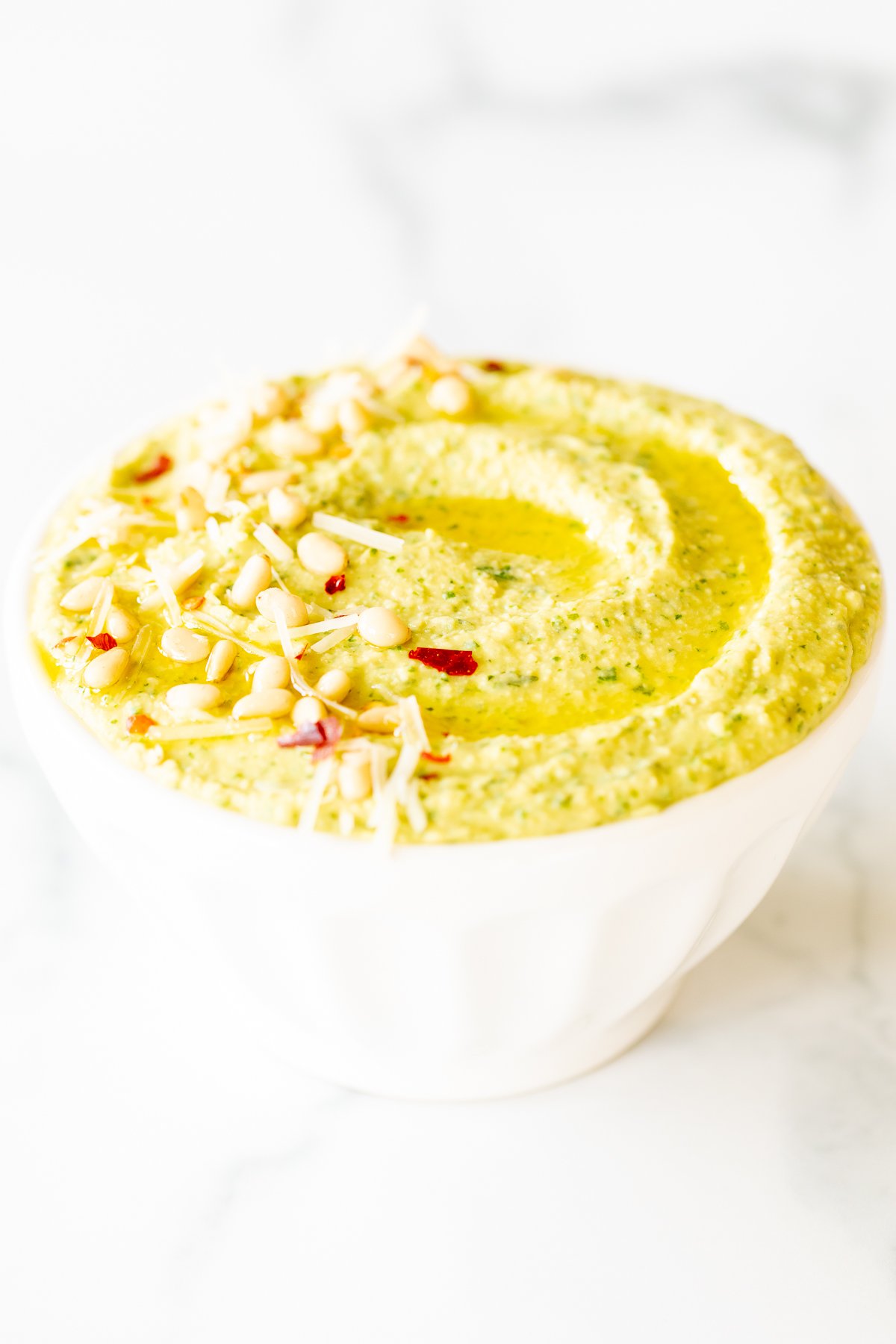 A white bowl full of basil pesto hummus, topped with pine nuts and parmesan.