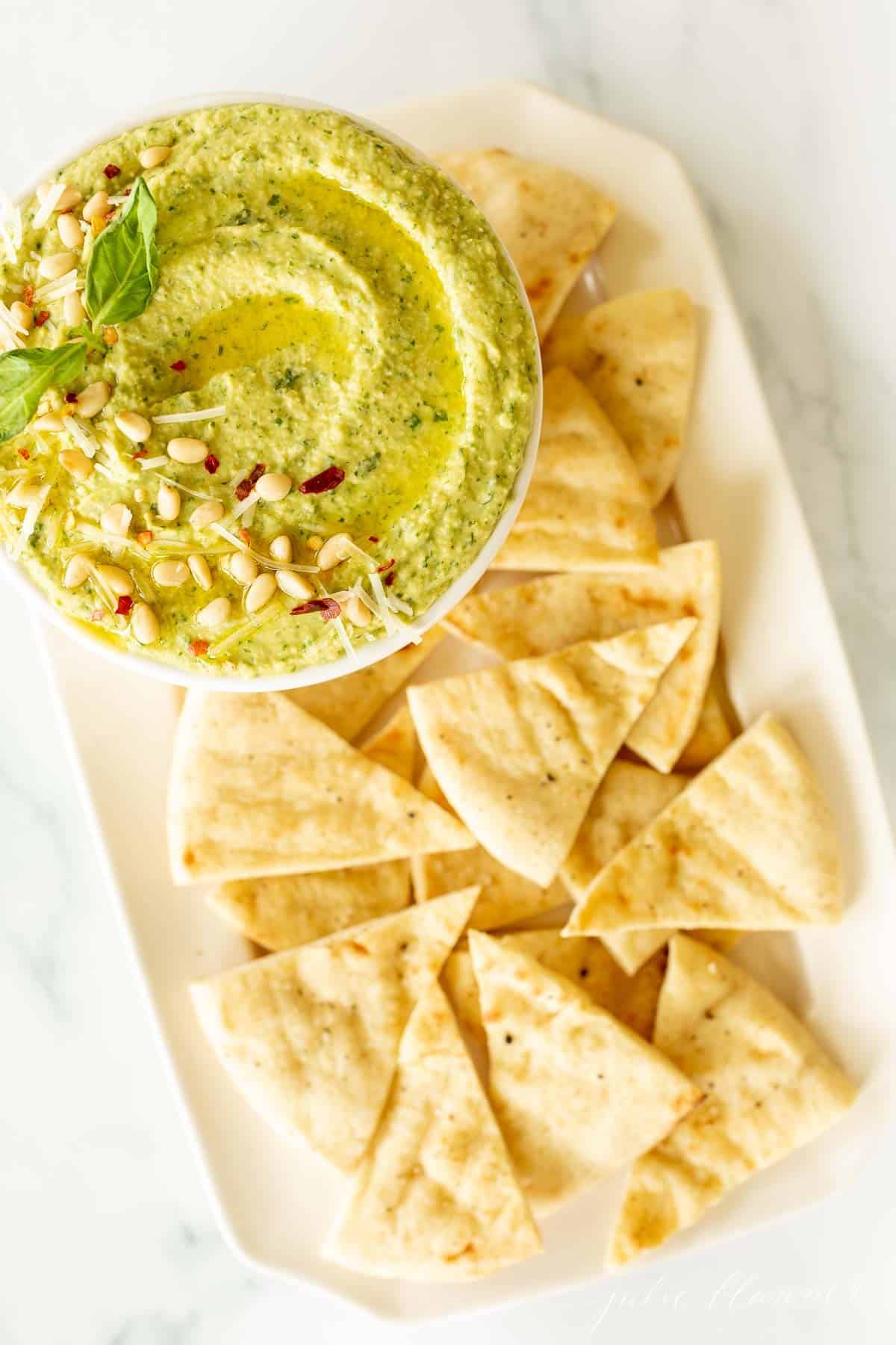 A platter full of pita triangles with a bowl full of basil pesto hummus.
