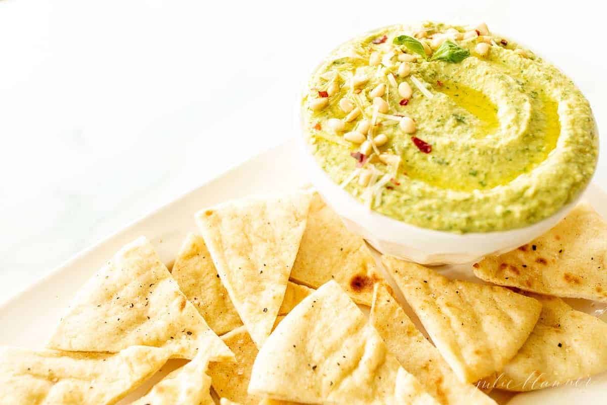 A platter full of pita triangles with a bowl full of basil pesto hummus.
