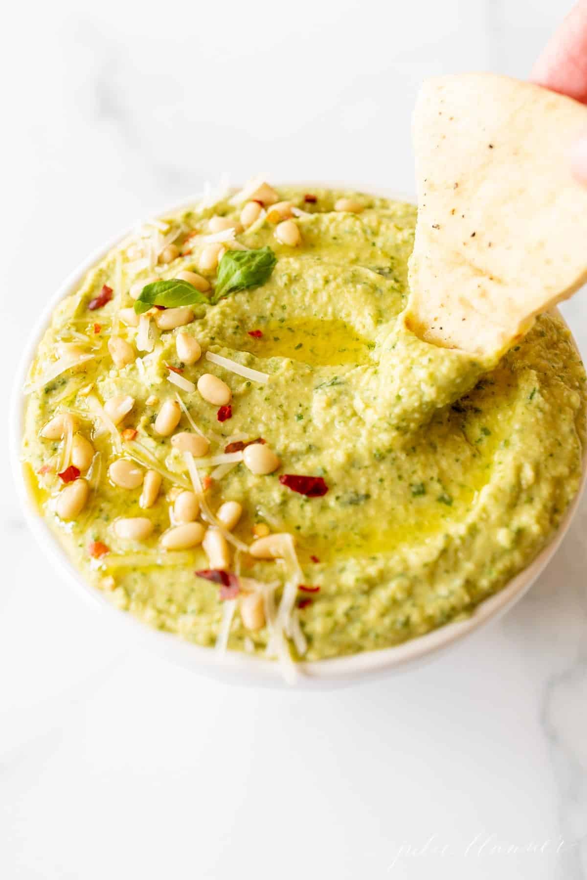 A bowl full of basil hummus on a marble surface, hand reaching in with a pita triangle.