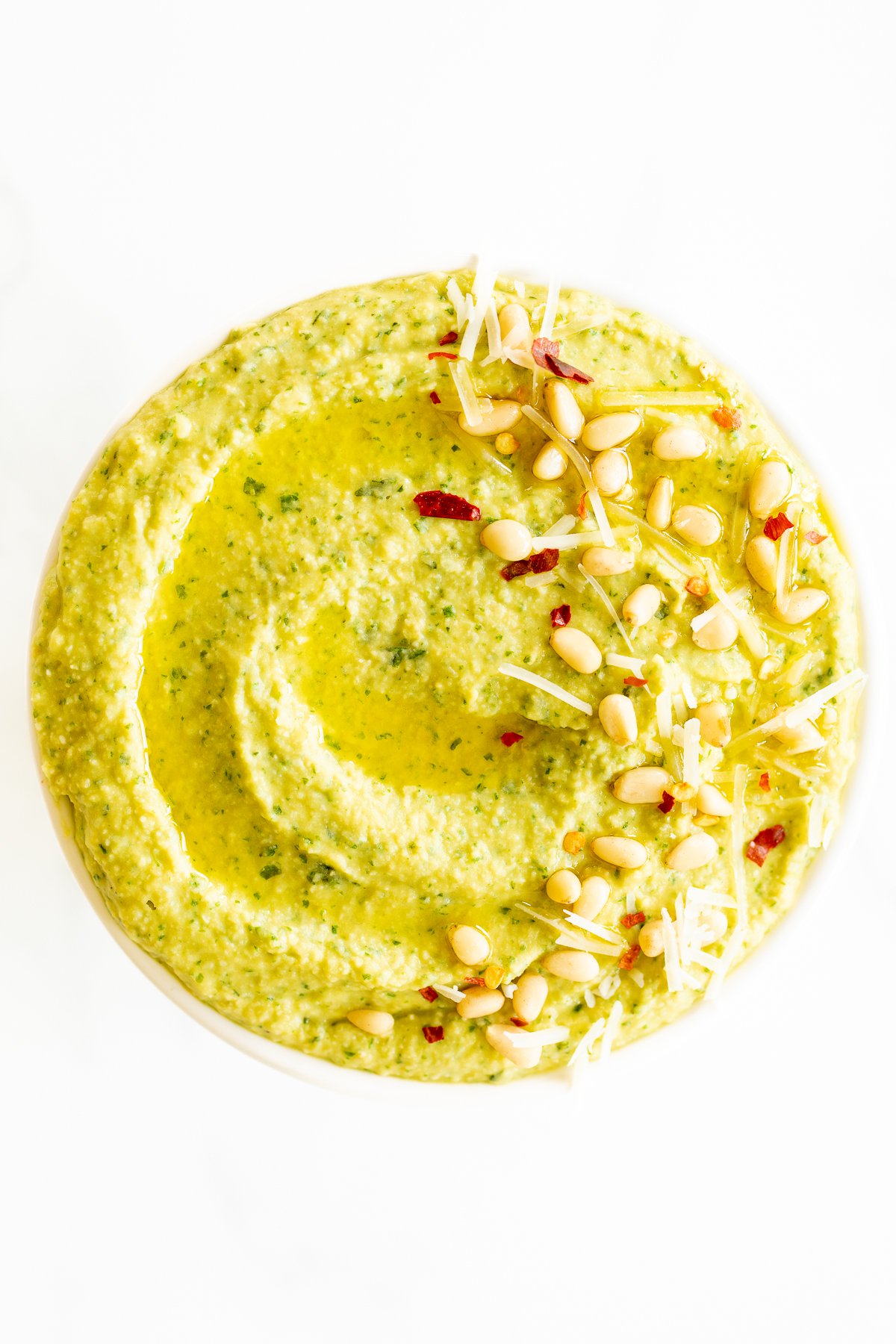 A white bowl full of basil pesto hummus, topped with pine nuts and parmesan.