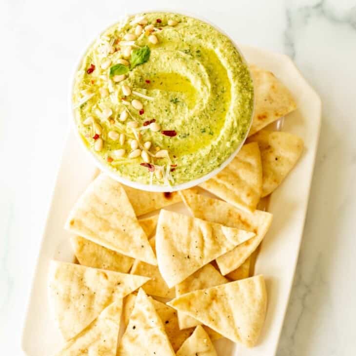 A platter full of pita triangles with a bowl full of basil pesto hummus.