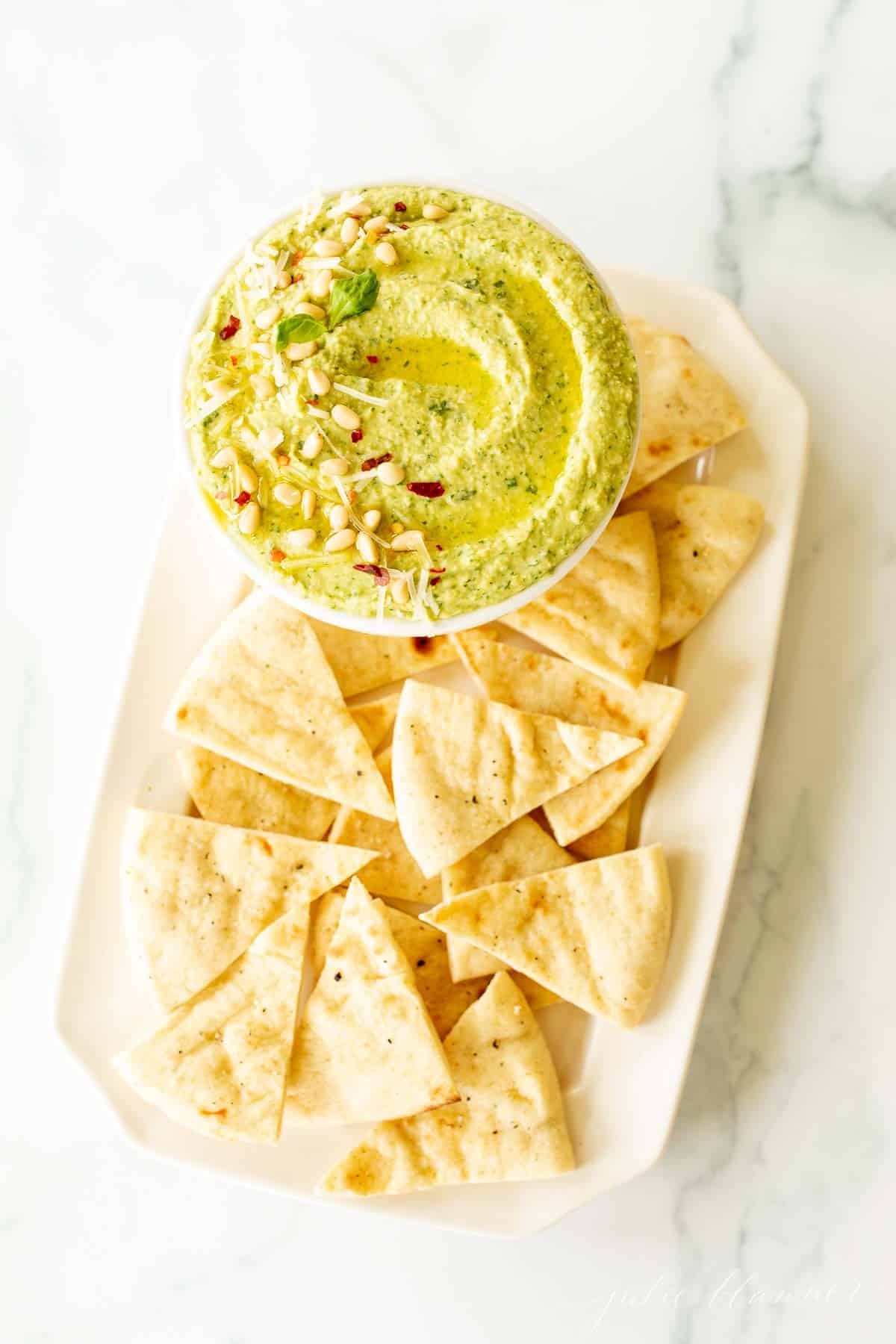 A platter full of pita triangles with a bowl full of basil pesto hummus.