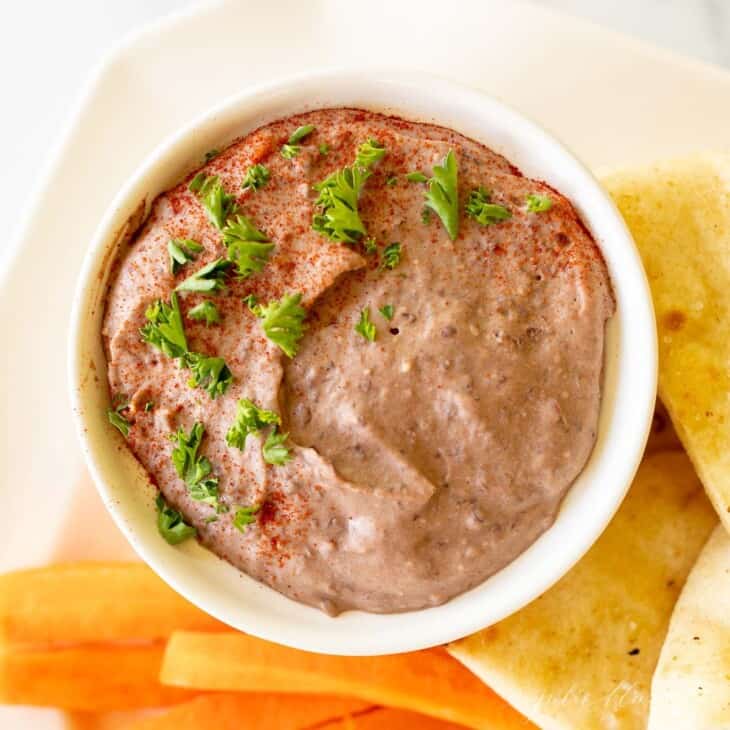 A white platter with a bowl of black bean hummus and pita triangles, celery and carrots.