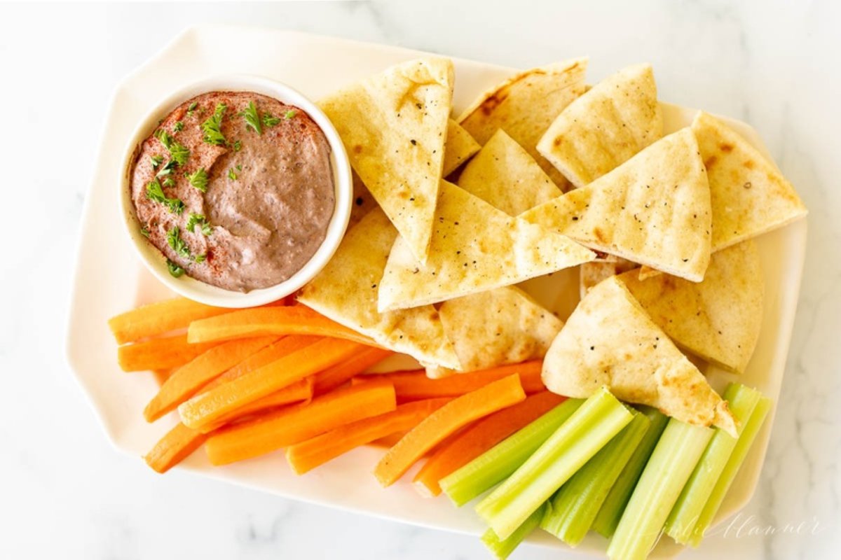 A white platter with pita slices, cut carrots and celery, with a small white bowl of black bean hummus.