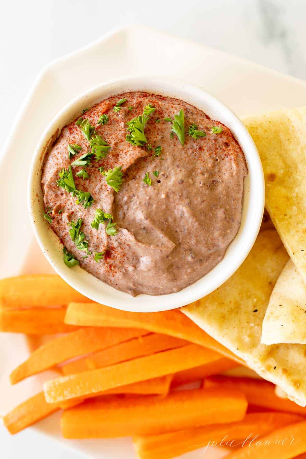 A white platter with a bowl of black bean hummus and pita triangles, celery and carrots.