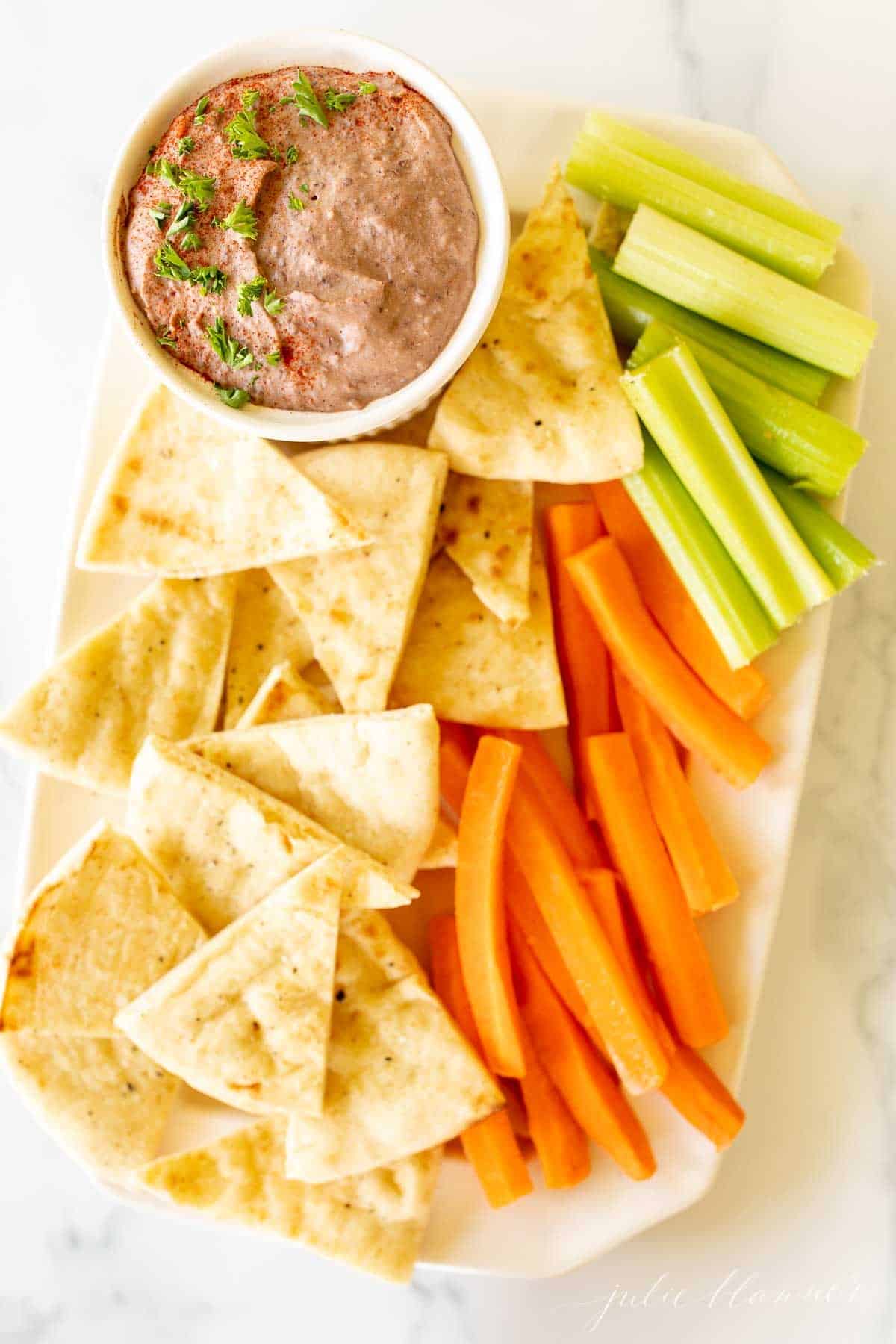 A white platter with a bowl of black bean hummus and pita triangles, celery and carrots.