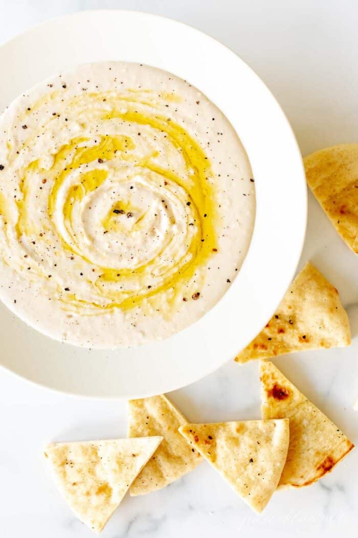 A white bowl filled with homemade white bean dip, drizzled with olive oil, pita triangles to the side.