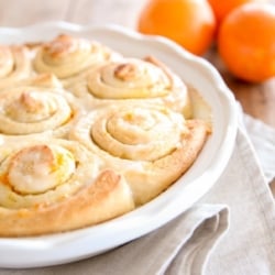 Orange rolls baked into a white ceramic round dish. Fresh oranges are on the table in the background.