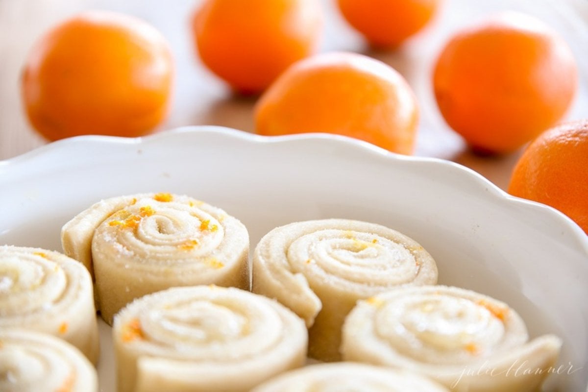 An orange rolls recipe, before baking, placed in a white ceramic round dish. Fresh oranges are on the table in the background.